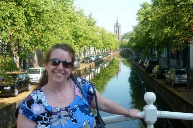Trudi standing on a bridge over a canal on a beautiful sunny day. She is smiling and wearing sun glasses.