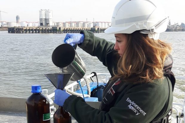 Clare standing next to the river, pouring from a metal jug into a glass bottle. She is wearing a white hard hat and an environment agency jacket