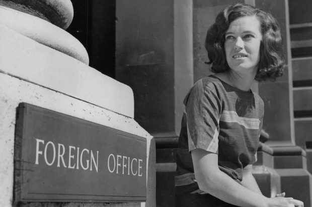 Black and white photograph of a woman sitting next to a sign which says Foreign Office.  The picture appears to be from the 1960s or 70s