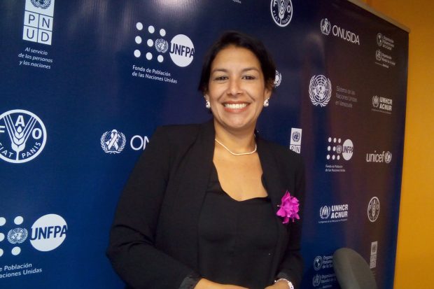 Picture of Irene in front of a blue banner displaying the names and logos of organisations. The organisations visible are PNUD, FAO, UNFPA, ONUSDA, UNHCR and Unicef