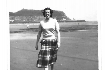 Black and white photo of Ellie's grandmother walking on the beach. She is wearing a pale jumper and tartan skirt. 