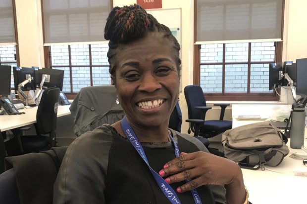 Tola smiling and sitting at her work desk. She is wearing a black top with a purple lanyard and is holding her hand to her chest.