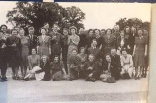 An old, black and white photograph of a group of 30-or-so people in 1940's clothing posing and smiling. Some are crouched or sat on the ground whilst the others stand behind them. The photo was taken outside on a lawn and there are tree-tops in the background.