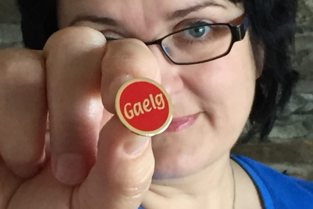 Picture of Breesha holding a badge up to the camera. The badge covers most of her face. It is red with the word "Gaelg" written in gold in the middle.