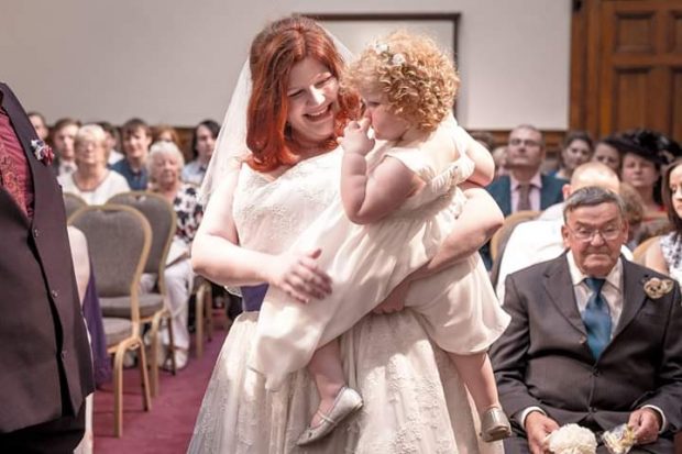 Photo of Rebeckah on her wedding day, standing at the front of a congregation and holding a little girl with blonde curly hair. Rebeckah is wearing a white wedding dress and veil.