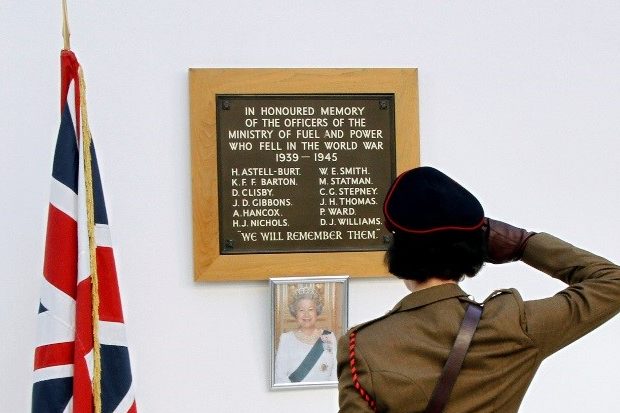 Photo of Kim saluting the British Flag, a picture of the queen and a plaque honouring those who fell during the second world war. The plaque reads "In honoured memory of the officers of the Ministry of Fuel and Power who fell in the World War 1939-1945. H. Astell-Burt, K.F.F Barton, D Clisby, J.D Gibbons, A Hancox, H.J Nichols, W.E Smith, M. Statman, C.G Stepney, J.H Thomas, P Ward, D.J Williams. We will remember them"