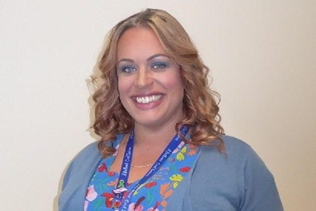 A head and shoulders shot of Emma, who has shoulder-length curly blonde hair, is smiling broadly and is wearing a blue flowery blouse.