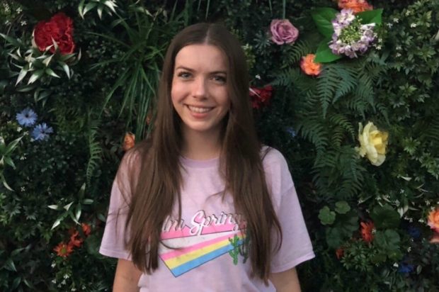 Sarah, a young-looking lady with long dark hair wears a pink t-shirt and smiles at the camera. In the background there are flowers and foliage.