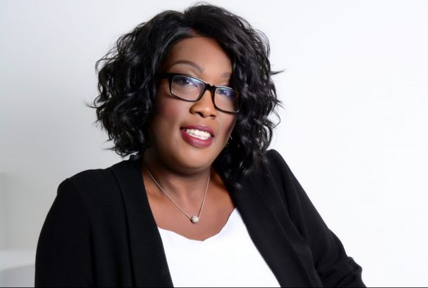 Picture of Cherron who is sitting in front of a white background. She is wearing glasses, a black jacket and a white top.